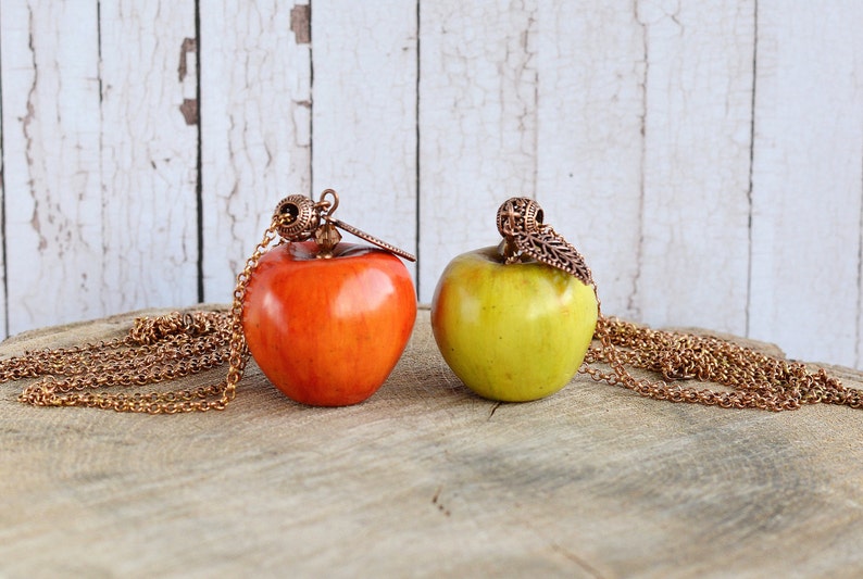 May include: Two apple-shaped pendants on gold chains. The apple on the left is red and the apple on the right is green.