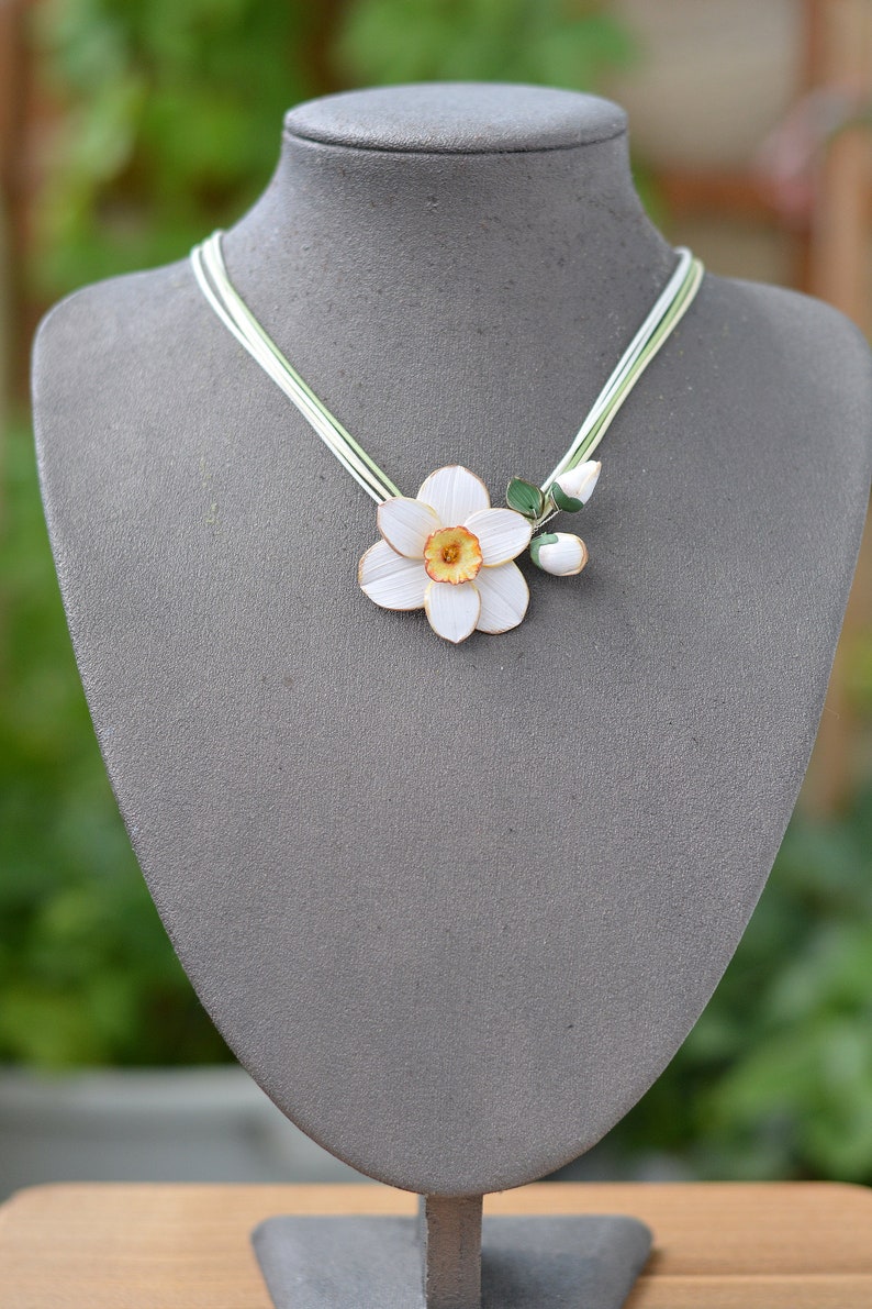 May include: A white and yellow paper flower necklace with green leaves. The necklace is on a grey mannequin bust.
