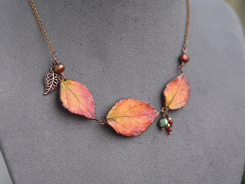 May include: A copper chain necklace with three dried leaves, a small copper leaf charm, and red and green beads.