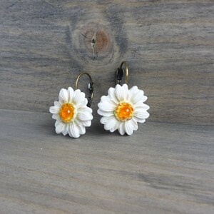 Op de afbeelding: Een paar madeliefjesoorbellen. Elke oorbel heeft een witte madeliefbloem met een feloranje centrum. De oorbellen hebben een klapsluiting en een bronzen metalen afwerking. De oorbellen staan tegen een houten achtergrond.