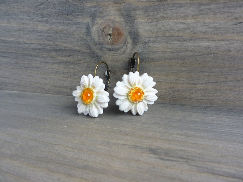 Op de afbeelding: Een paar madeliefjesoorbellen. Elke oorbel heeft een witte madeliefbloem met een feloranje centrum. De oorbellen hebben een klapsluiting en een bronzen metalen afwerking. De oorbellen staan tegen een houten achtergrond.