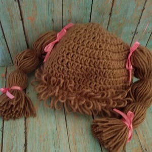 May include: A brown crocheted hat with attached yarn braids and tassels, resembling a doll's hairstyle. The hat is adorned with pink ribbons at the base of each braid and tassel. The hat is designed to be worn.
