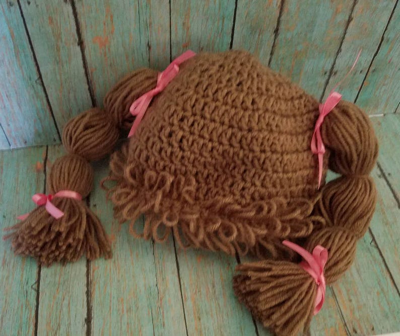 May include: A brown crocheted hat with attached yarn braids and tassels, resembling a doll's hairstyle. The hat is adorned with pink ribbons at the base of each braid and tassel. The hat is designed to be worn.
