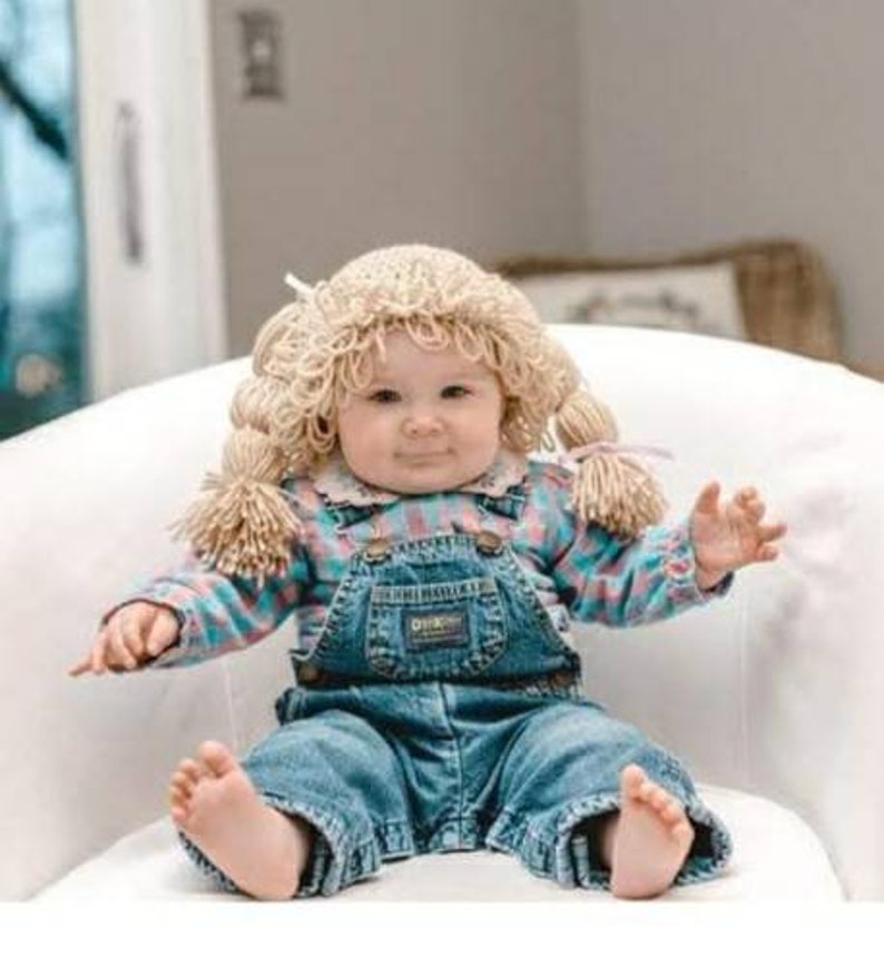 May include: A baby wearing a blonde yarn wig with pigtails, a blue, pink, and white plaid shirt, and denim overalls. The overalls have a front pocket and the word "OSH KOSH" on the bib.