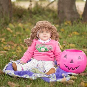 May include: A baby wearing a brown yarn wig and a pink sweatshirt with the text "Cabbage Patch Kids". The baby is sitting on a blanket next to a pink jack-o'-lantern bucket. The baby is wearing white shoes and floral pants.