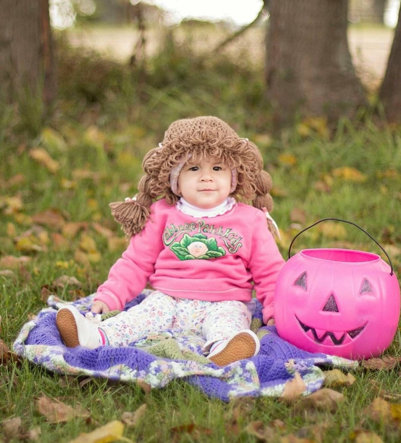 May include: A baby wearing a brown yarn wig and a pink sweatshirt with the text "Cabbage Patch Kids". The baby is sitting on a blanket next to a pink jack-o'-lantern bucket. The baby is wearing white shoes and floral pants.
