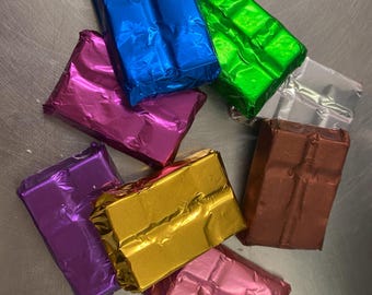 May include: An assortment of individually wrapped rectangular items in various colors, including blue, green, silver, brown, gold, pink, and purple. The shiny, metallic wrapping paper gives a festive appearance. The items are arranged on a metallic surface.