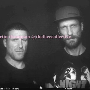 May include: Two men, one wearing a black shirt and the other wearing a black shirt with a graphic print of the word "NIGHT" and a person's face, are standing side by side. The man on the right is wearing a black baseball cap with a white logo.