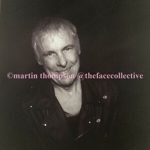 May include: A black and white photo of a man with short, gray hair. He is smiling and wearing a black leather jacket. The photo is taken from the chest up and the background is dark.