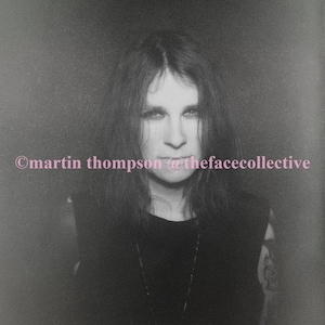 May include: A black and white portrait of a person with long dark hair. The person is wearing a black shirt with a silver necklace. The image is grainy and has a vintage feel.