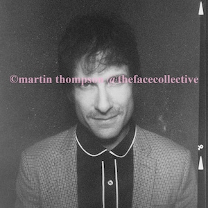 May include: A black and white photo of a person wearing a checkered blazer over a black collared shirt with white piping. The person is smiling and looking at the camera. The photo has the text "©martin thompson @thefacecollective" at the top.