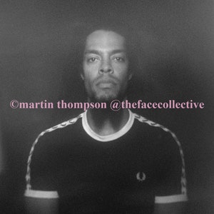 May include: A black and white photo of a person wearing a black t-shirt with a white collar and white stripes on the sleeves. The t-shirt has a small white logo on the chest. The person is looking at the camera. ©martin thompson @thefacecollective