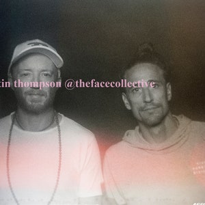 May include: Two men are standing side-by-side, looking at the camera. The man on the left is wearing a white baseball cap, a white t-shirt, and a beaded necklace. The man on the right is wearing a light brown hoodie. The photo is in black and white and has a grainy, vintage filter applied.