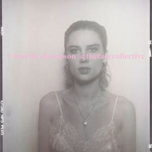May include: A black and white photo of a person wearing a white lace top and a silver necklace with a pendant. The photo is taken in a photo booth and has the text "@thefacecollective" printed on it.