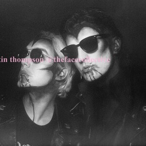 May include: Two people wearing sunglasses and black leather jackets with makeup on their faces. The person on the left has a black and white design on their face and the person on the right has a red design on their face. The text "©martin thompson & thefacecollective" is in the bottom left corner of the image.