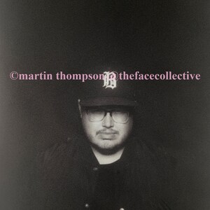 May include: A black and white photograph of a person wearing a black baseball cap with a white letter "B" on the front, black framed glasses, and a black shirt. The text "©martin thompson®thefacecollective" is at the top of the image.