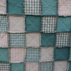 May include: A patchwork quilt with squares of green and beige fabric. The fabric has different patterns, including plaid, stripes, and a simple diamond pattern. The quilt is made with a frayed edge.
