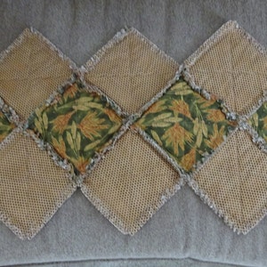 K&ouml;nnte beinhalten: Ein Tischl&auml;ufer aus Patchwork-Stoffquadraten. Die Quadrate sind eine Mischung aus Braun und Gr&uuml;n mit einem Blumenmuster. Der L&auml;ufer ist an allen Kanten gefranstet.