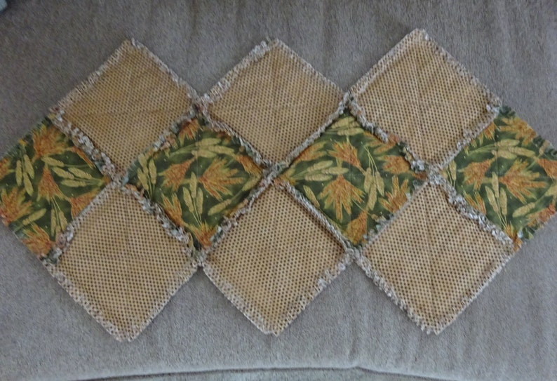 K&ouml;nnte beinhalten: Ein Tischl&auml;ufer aus Patchwork-Stoffquadraten. Die Quadrate sind eine Mischung aus Braun und Gr&uuml;n mit einem Blumenmuster. Der L&auml;ufer ist an allen Kanten gefranstet.