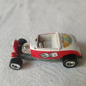 May include: A red and white toy hot rod car featuring a graphic of two dice on the side panel and a fan of playing cards on the rear deck. The vehicle has an exposed silver engine, black wheels with chrome rims, and a white interior with a steering wheel.