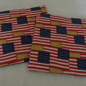 May include: Two red, white, and blue American flag patterned pot holders. The pot holders are quilted and have a tan border.