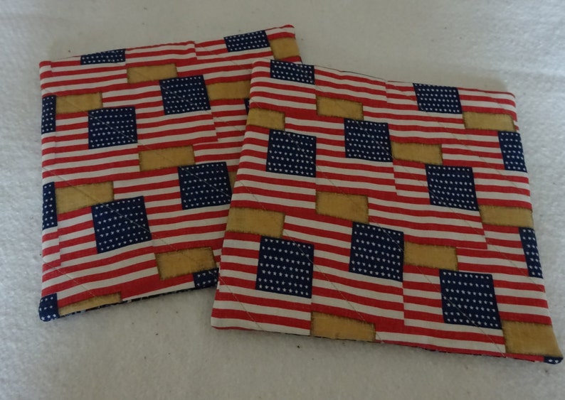 May include: Two red, white, and blue American flag patterned pot holders. The pot holders are quilted and have a tan border.