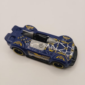 May include: A dark blue toy race car featuring yellow geometric accents and silver detailing on the hood and cockpit area. The vehicle has black wheels with yellow rims, a low-profile design, and a small silver fin behind the open driver seat.