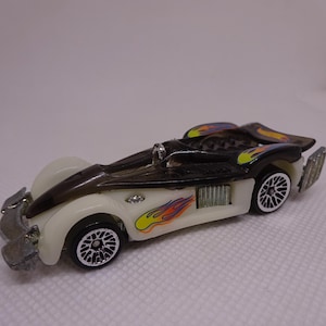 May include: A toy car with a sleek design, featuring a white body with black and transparent accents. The car has flame decals in yellow, orange, and red, and black wheels with silver rims. The car's front and rear have silver details.