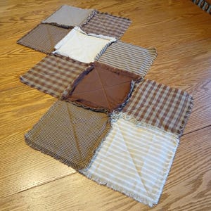 May include: A patchwork quilt composed of nine squares in various brown and cream patterns, including stripes, checks, and solid fabrics. The quilt has a fringed edge and is arranged diagonally on a wooden surface.