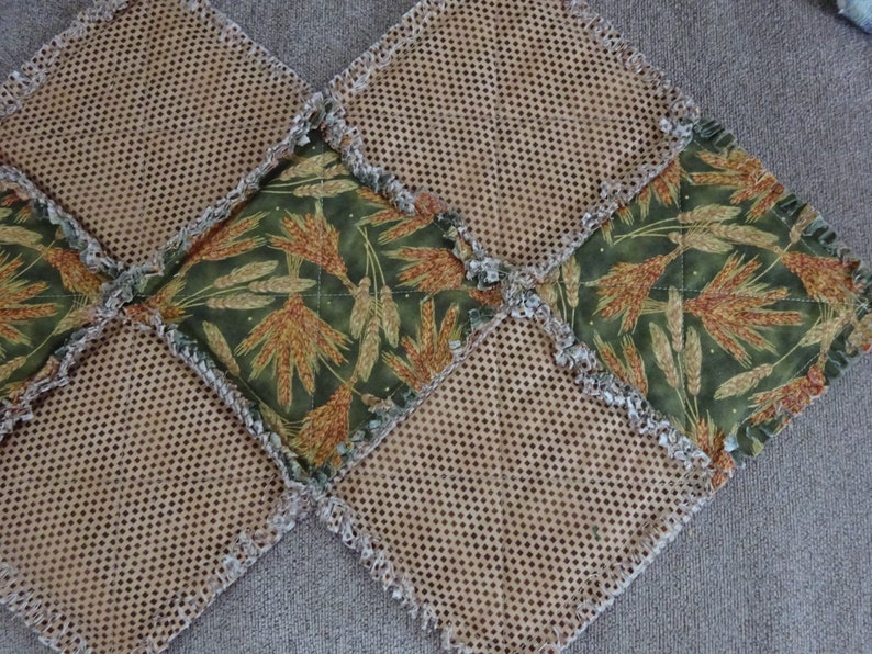 K&ouml;nnte beinhalten: Eine braune und gr&uuml;ne Patchwork-Decke mit ausgefransten Kanten. Die Decke besteht aus Quadraten aus verschiedenen Stoffen, darunter ein braunes Netzgewebe und ein gr&uuml;ner Stoff mit einem Weizenmuster.
