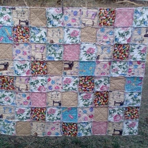 May include: A patchwork quilt with a variety of floral and sewing-themed fabrics. The quilt is made up of squares of different colors and patterns, including pink, blue, green, and brown. The quilt is hanging on a clothesline.