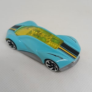 May include: A light blue toy car with a yellow translucent canopy, featuring black and yellow racing stripes along the top. The vehicle has a grey lower chassis and black wheels with white rims, displaying the Hot Wheels logo on the rear fender.