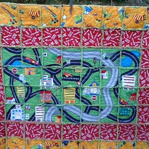 May include: A patchwork quilt with a city road theme. The quilt is made of squares of fabric with different designs, including cars, trucks, buildings, and stop signs. The quilt is edged with a green fringe.