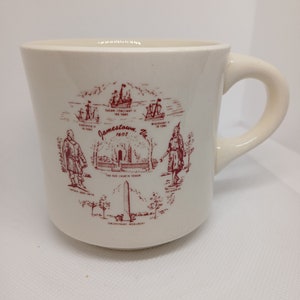 May include: Cream-colored ceramic mug with a curved handle. The mug features a maroon illustration of Jamestown, Virginia, including ships, historical figures, and the text "Jamestown 1607".