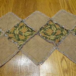 K&ouml;nnte beinhalten: Ein brauner und gr&uuml;ner Tischl&auml;ufer mit Patchwork-Muster. Der L&auml;ufer besteht aus Quadraten aus braunem Netzstoff und gr&uuml;nem Stoff mit einem Muster aus Weizen&auml;hren. Der L&auml;ufer hat einen ausgefransten Rand.