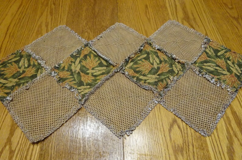 K&ouml;nnte beinhalten: Ein brauner und gr&uuml;ner Tischl&auml;ufer mit Patchwork-Muster. Der L&auml;ufer besteht aus Quadraten aus braunem Netzstoff und gr&uuml;nem Stoff mit einem Muster aus Weizen&auml;hren. Der L&auml;ufer hat einen ausgefransten Rand.