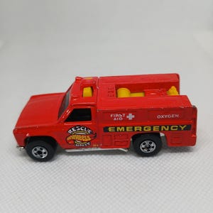 May include: A red Hot Wheels Rescue Wheels Unit toy truck. The toy truck has black wheels and features the words "EMERGENCY", "FIRST AID", and "OXYGEN". The truck has yellow cylinders on the back.