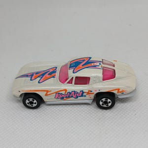 May include: A white toy car with a stylized design in blue, pink, and orange. The car has the text "Kool-Aid" on the side. The car has black wheels and a sleek, aerodynamic shape.