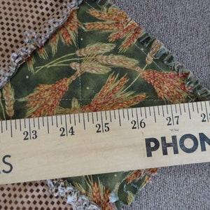 K&ouml;nnte beinhalten: Nahaufnahme eines Holzlineals mit den aufgedruckten W&ouml;rtern "PHONE" und "PLIES". Das Lineal misst ein St&uuml;ck Stoff mit einem gr&uuml;nen und braunen Weizenmuster.