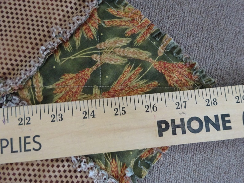 K&ouml;nnte beinhalten: Nahaufnahme eines Holzlineals mit den aufgedruckten W&ouml;rtern "PHONE" und "PLIES". Das Lineal misst ein St&uuml;ck Stoff mit einem gr&uuml;nen und braunen Weizenmuster.