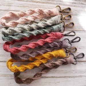 May include: A collection of six macrame wristlet keychains in various colors including light pink, sage green, dusty rose, dark gray, mustard yellow, and brown. Each keychain features a twisted cord design and a metal lobster clasp.