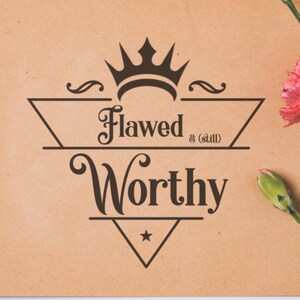 Op de afbeelding: Een bruine kaart met de tekst "Flawed & (still) Worthy" in een decoratief lettertype, bekroond met een kroonontwerp. Roze anjers en een bloemknop bevinden zich in de rechterbovenhoek.