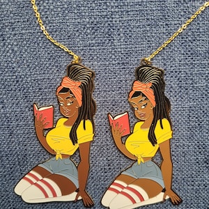 May include: Two gold-toned necklaces each feature a pendant of a woman reading a book. The woman is depicted in a yellow top, denim shorts, and knee-high socks with red stripes. The pendants are on a blue denim background.