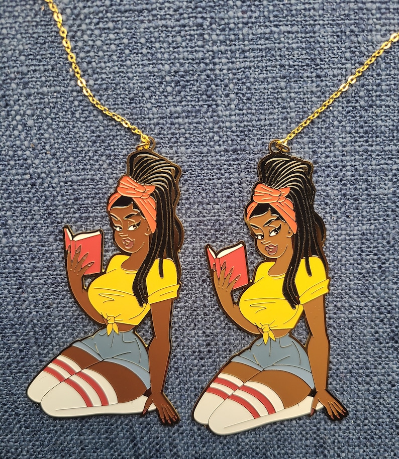 May include: Two gold-toned necklaces each feature a pendant of a woman reading a book. The woman is depicted in a yellow top, denim shorts, and knee-high socks with red stripes. The pendants are on a blue denim background.