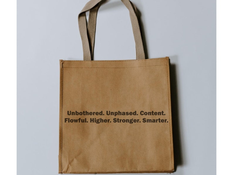 May include: A brown paper tote bag with beige fabric handles. The bag has the text "Unbothered. Unphased. Content. Flowful. Higher. Stronger. Smarter." printed on the front.