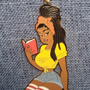 May include: A gold-colored pendant featuring a cartoon illustration of a woman reading a book. The woman is wearing a yellow top, denim shorts, and knee-high socks. Her hair is styled with dreadlocks and an orange headscarf. The pendant is on a gold chain.