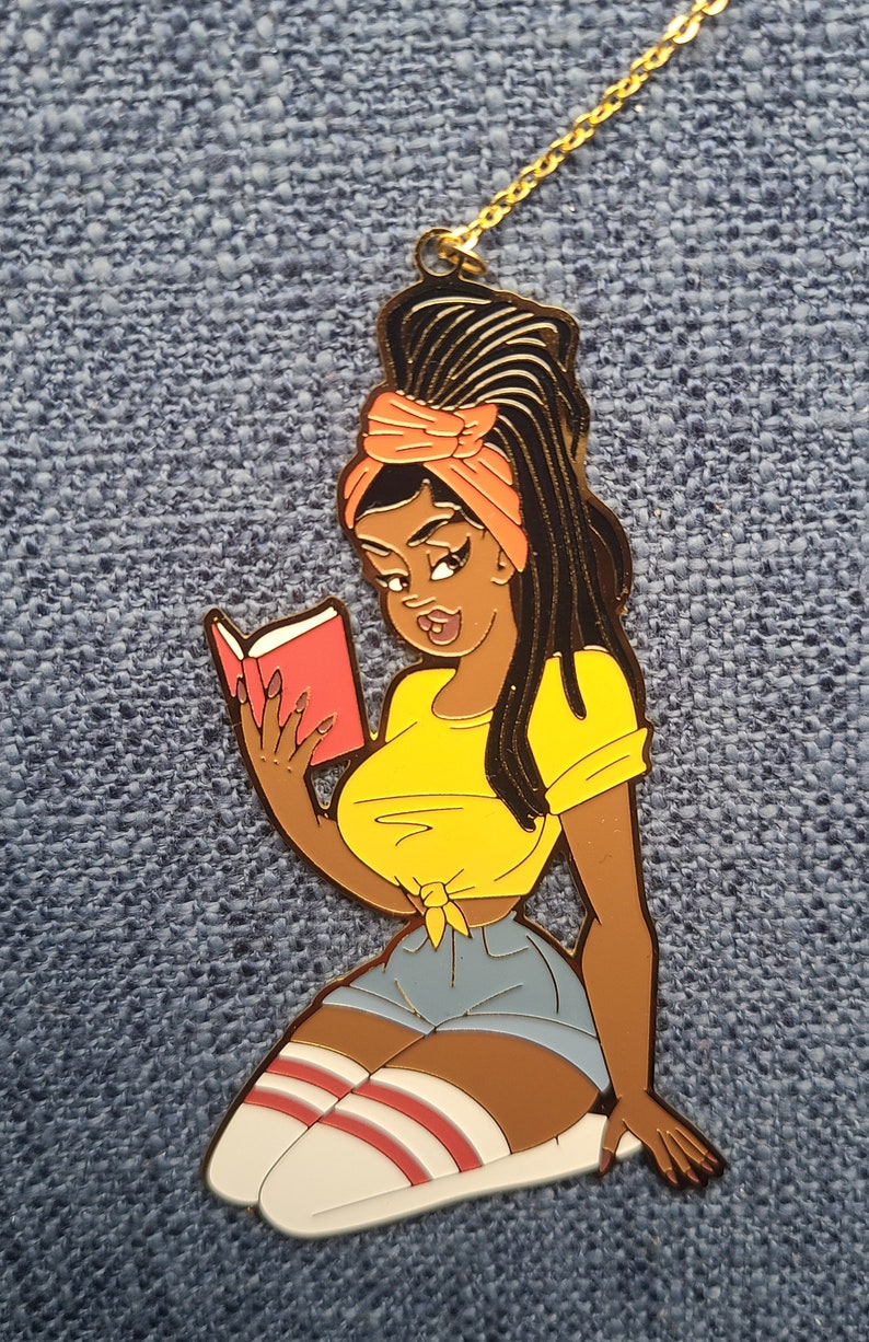 May include: A gold-colored pendant featuring a cartoon illustration of a woman reading a book. The woman is wearing a yellow top, denim shorts, and knee-high socks. Her hair is styled with dreadlocks and an orange headscarf. The pendant is on a gold chain.