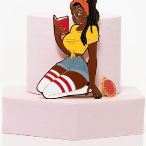 May include: A metal pin of a Black woman reading a book, wearing a yellow top, denim shorts, and knee-high socks with red stripes. The pin has a gold-colored loop at the top and a small charm that says "The Book Babes".