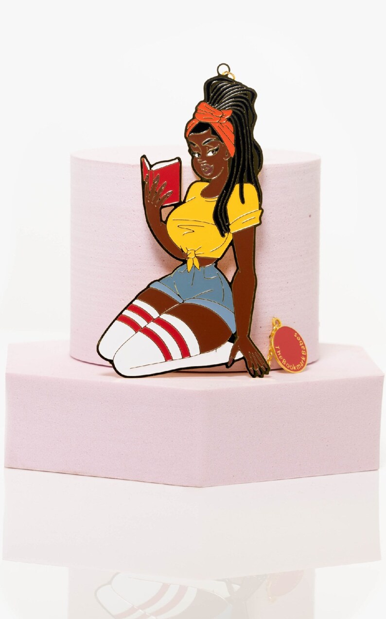 May include: A metal pin of a Black woman reading a book, wearing a yellow top, denim shorts, and knee-high socks with red stripes. The pin has a gold-colored loop at the top and a small charm that says "The Book Babes".