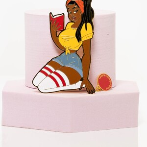 May include: A colorful enamel pin featuring a cartoon woman reading a book, wearing a yellow top, denim shorts, and knee-high socks with red stripes. A smaller pin with the text "The Bookmark Babes" is also present.